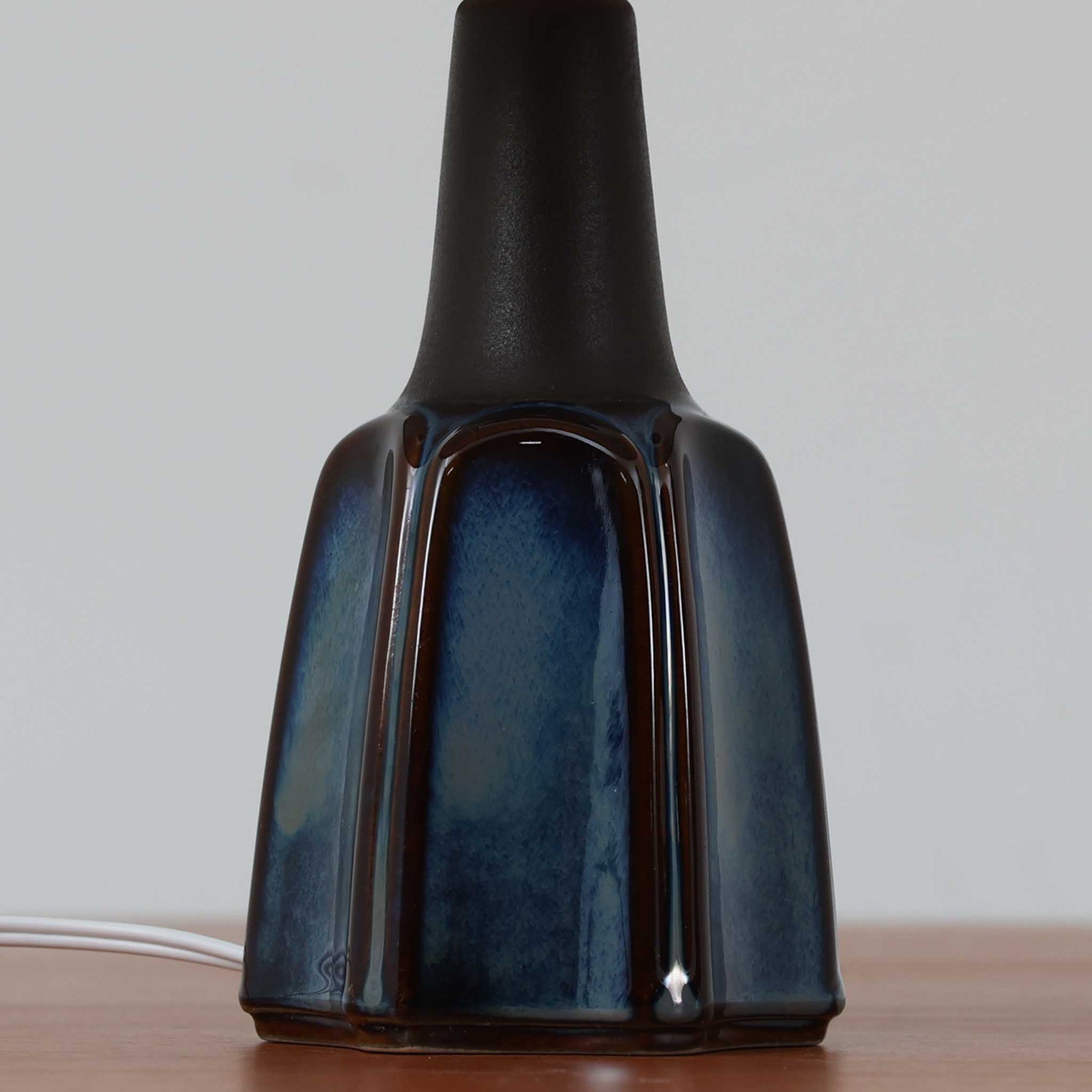 Einar JohansenがSoholm Keramik（スーホルム・セラミック）のためにデザインしたモデル1031は、深みのあるブルーの釉薬と立体的な面取りが織りなす陰影が美しい陶磁器ベースが特徴で、上部のマットな質感との対比がモダンで彫刻的な存在感を放つ北欧ビンテージ品です。プリーツシェードを透ける柔らかな灯りが落ち着いた雰囲気をつくり、ベッドサイドやリビングのアクセント照明として取り入れやすく、経年で深まった釉薬の表情が一点物ならではの魅力を一層引き立てています。