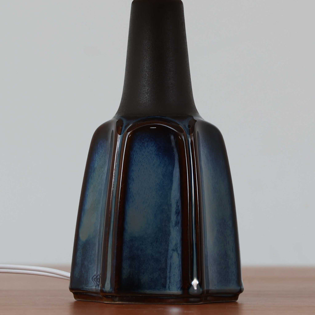 Einar JohansenがSoholm Keramik（スーホルム・セラミック）のためにデザインしたモデル1031は、深みのあるブルーの釉薬と立体的な面取りが織りなす陰影が美しい陶磁器ベースが特徴で、上部のマットな質感との対比がモダンで彫刻的な存在感を放つ北欧ビンテージ品です。プリーツシェードを透ける柔らかな灯りが落ち着いた雰囲気をつくり、ベッドサイドやリビングのアクセント照明として取り入れやすく、経年で深まった釉薬の表情が一点物ならではの魅力を一層引き立てています。