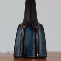 Einar JohansenがSoholm Keramik（スーホルム・セラミック）のためにデザインしたモデル1031は、深みのあるブルーの釉薬と立体的な面取りが織りなす陰影が美しい陶磁器ベースが特徴で、上部のマットな質感との対比がモダンで彫刻的な存在感を放つ北欧ビンテージ品です。プリーツシェードを透ける柔らかな灯りが落ち着いた雰囲気をつくり、ベッドサイドやリビングのアクセント照明として取り入れやすく、経年で深まった釉薬の表情が一点物ならではの魅力を一層引き立てています。