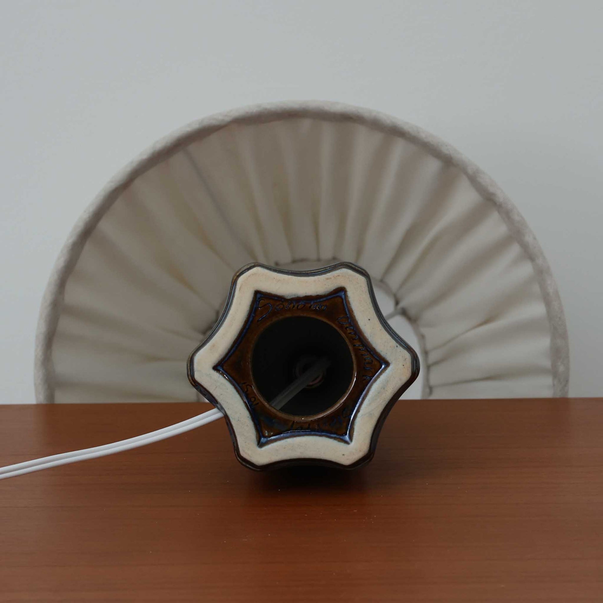 Einar JohansenがSoholm Keramik（スーホルム・セラミック）のためにデザインしたモデル1031は、深みのあるブルーの釉薬と立体的な面取りが織りなす陰影が美しい陶磁器ベースが特徴で、上部のマットな質感との対比がモダンで彫刻的な存在感を放つ北欧ビンテージ品です。プリーツシェードを透ける柔らかな灯りが落ち着いた雰囲気をつくり、ベッドサイドやリビングのアクセント照明として取り入れやすく、経年で深まった釉薬の表情が一点物ならではの魅力を一層引き立てています。