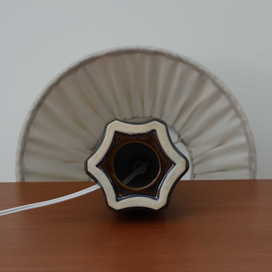 Einar JohansenがSoholm Keramik（スーホルム・セラミック）のためにデザインしたモデル1031は、深みのあるブルーの釉薬と立体的な面取りが織りなす陰影が美しい陶磁器ベースが特徴で、上部のマットな質感との対比がモダンで彫刻的な存在感を放つ北欧ビンテージ品です。プリーツシェードを透ける柔らかな灯りが落ち着いた雰囲気をつくり、ベッドサイドやリビングのアクセント照明として取り入れやすく、経年で深まった釉薬の表情が一点物ならではの魅力を一層引き立てています。