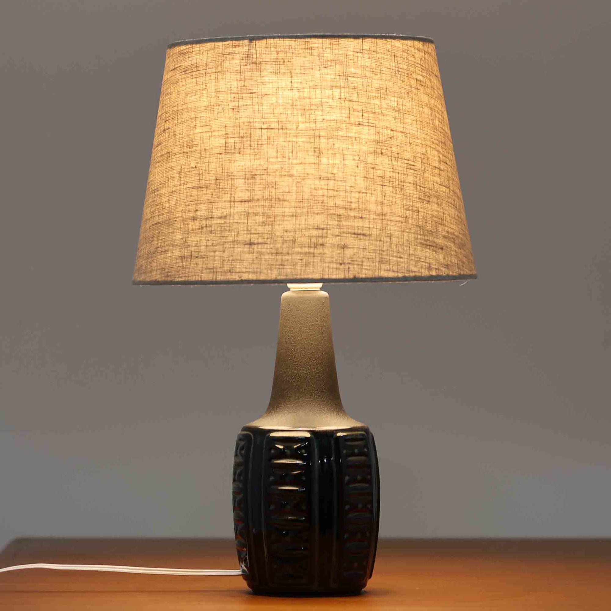 Einar JohansenがSoholm Keramik（スーホルム・セラミック）のために手掛けたモデル1038は、深みのあるブルーの釉薬が重なり合うように陰影をつくる陶磁器ベースと、上部のマットな質感とのコントラストが美しい北欧ビンテージ品で、四面に施された力強いレリーフ装飾が彫刻的な存在感を与えます。リネンシェード越しに広がる柔らかな灯りが空間を優しく包み込み、リビングやベッドサイドのアクセント照明として取り入れやすく、経年で深まった釉薬の表情が一点物ならではの味わいを一層引き立てています。