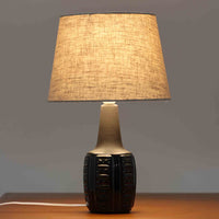 Einar JohansenがSoholm Keramik（スーホルム・セラミック）のために手掛けたモデル1038は、深みのあるブルーの釉薬が重なり合うように陰影をつくる陶磁器ベースと、上部のマットな質感とのコントラストが美しい北欧ビンテージ品で、四面に施された力強いレリーフ装飾が彫刻的な存在感を与えます。リネンシェード越しに広がる柔らかな灯りが空間を優しく包み込み、リビングやベッドサイドのアクセント照明として取り入れやすく、経年で深まった釉薬の表情が一点物ならではの味わいを一層引き立てています。