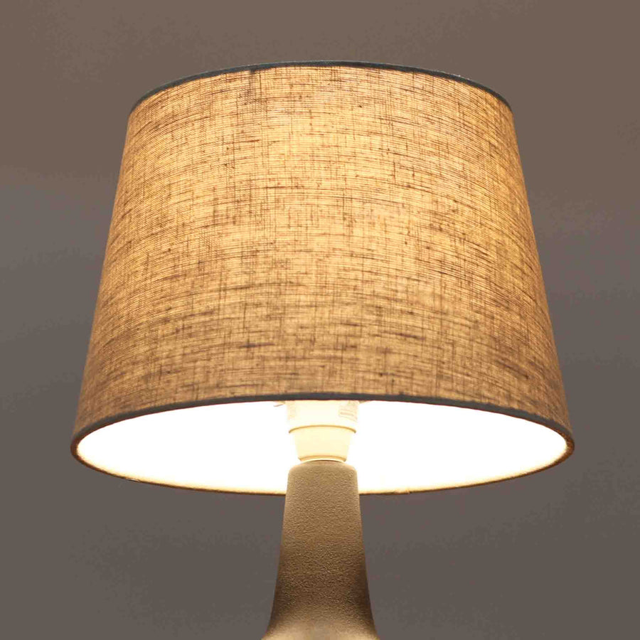 Einar JohansenがSoholm Keramik（スーホルム・セラミック）のために手掛けたモデル1038は、深みのあるブルーの釉薬が重なり合うように陰影をつくる陶磁器ベースと、上部のマットな質感とのコントラストが美しい北欧ビンテージ品で、四面に施された力強いレリーフ装飾が彫刻的な存在感を与えます。リネンシェード越しに広がる柔らかな灯りが空間を優しく包み込み、リビングやベッドサイドのアクセント照明として取り入れやすく、経年で深まった釉薬の表情が一点物ならではの味わいを一層引き立てています。