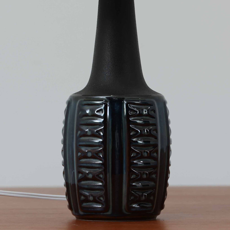 Einar JohansenがSoholm Keramik（スーホルム・セラミック）のために手掛けたモデル1038は、深みのあるブルーの釉薬が重なり合うように陰影をつくる陶磁器ベースと、上部のマットな質感とのコントラストが美しい北欧ビンテージ品で、四面に施された力強いレリーフ装飾が彫刻的な存在感を与えます。リネンシェード越しに広がる柔らかな灯りが空間を優しく包み込み、リビングやベッドサイドのアクセント照明として取り入れやすく、経年で深まった釉薬の表情が一点物ならではの味わいを一層引き立てています。