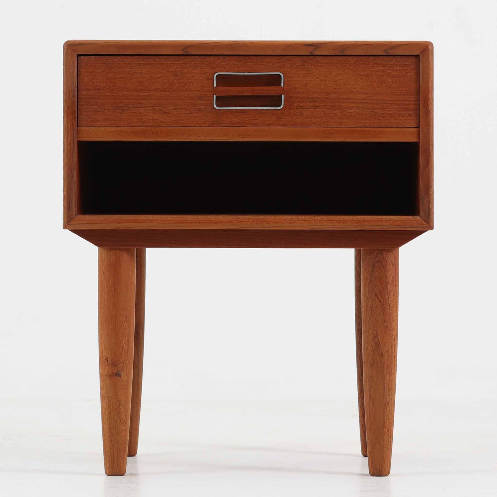 デンマーク製のサイドチェストで、シンプルなボックス構成にチーク材の豊かな風合いが映える北欧ビンテージらしい一台です。オープンスペースは本や小物の収納に便利で、引き出しには金属フレームを組み合わせたユニークな取手がアクセントとして効いています。丸みを帯びた角や細く伸びたテーパードレッグが軽快な印象を与え、ベッドサイドやソファ横でのサイドテーブル使いにも最適。経年変化の味わい深い木肌が空間に温かさと上質さを添えます。