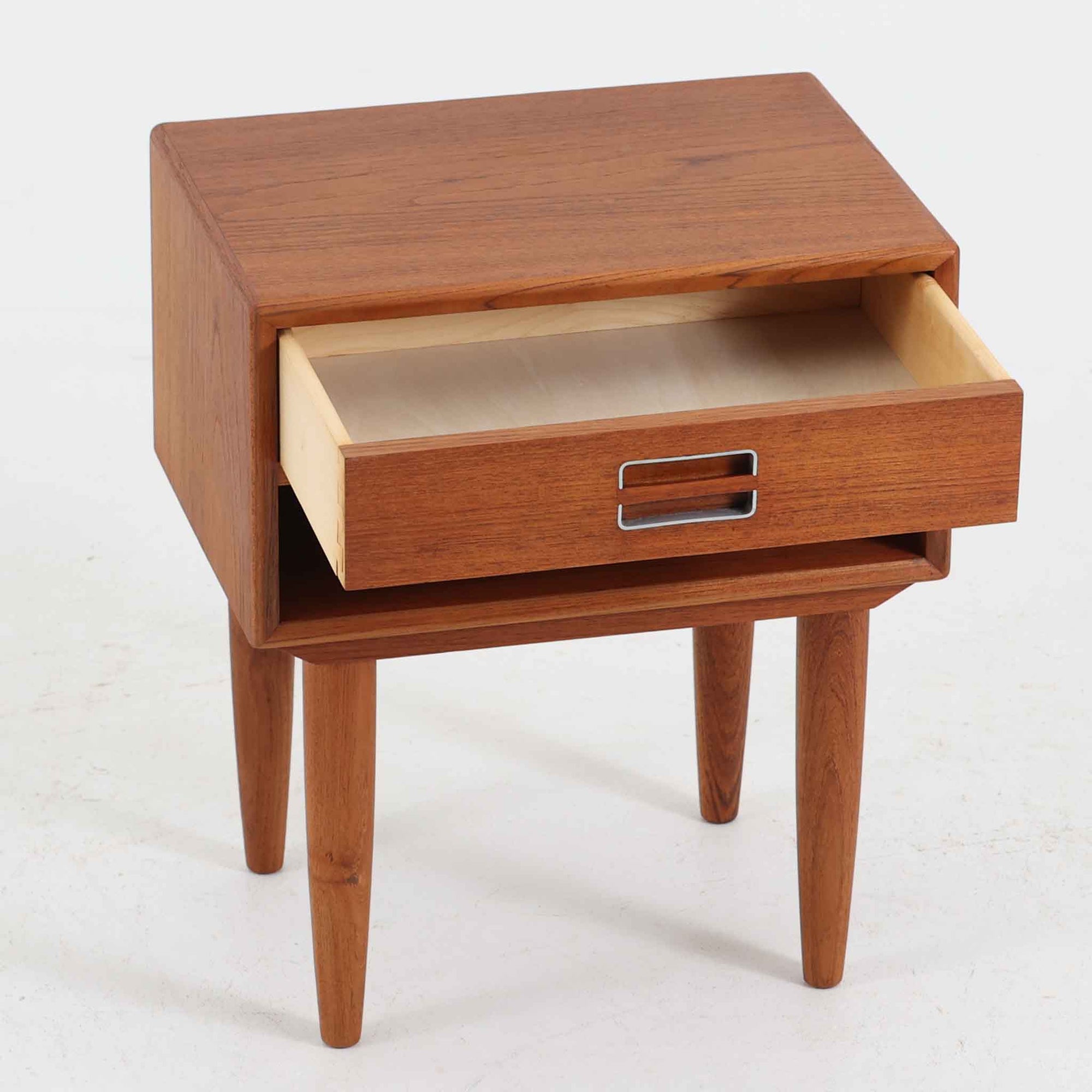 デンマーク製のサイドチェストで、シンプルなボックス構成にチーク材の豊かな風合いが映える北欧ビンテージらしい一台です。オープンスペースは本や小物の収納に便利で、引き出しには金属フレームを組み合わせたユニークな取手がアクセントとして効いています。丸みを帯びた角や細く伸びたテーパードレッグが軽快な印象を与え、ベッドサイドやソファ横でのサイドテーブル使いにも最適。経年変化の味わい深い木肌が空間に温かさと上質さを添えます。