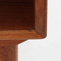 デンマーク製のサイドチェストで、シンプルなボックス構成にチーク材の豊かな風合いが映える北欧ビンテージらしい一台です。オープンスペースは本や小物の収納に便利で、引き出しには金属フレームを組み合わせたユニークな取手がアクセントとして効いています。丸みを帯びた角や細く伸びたテーパードレッグが軽快な印象を与え、ベッドサイドやソファ横でのサイドテーブル使いにも最適。経年変化の味わい深い木肌が空間に温かさと上質さを添えます。