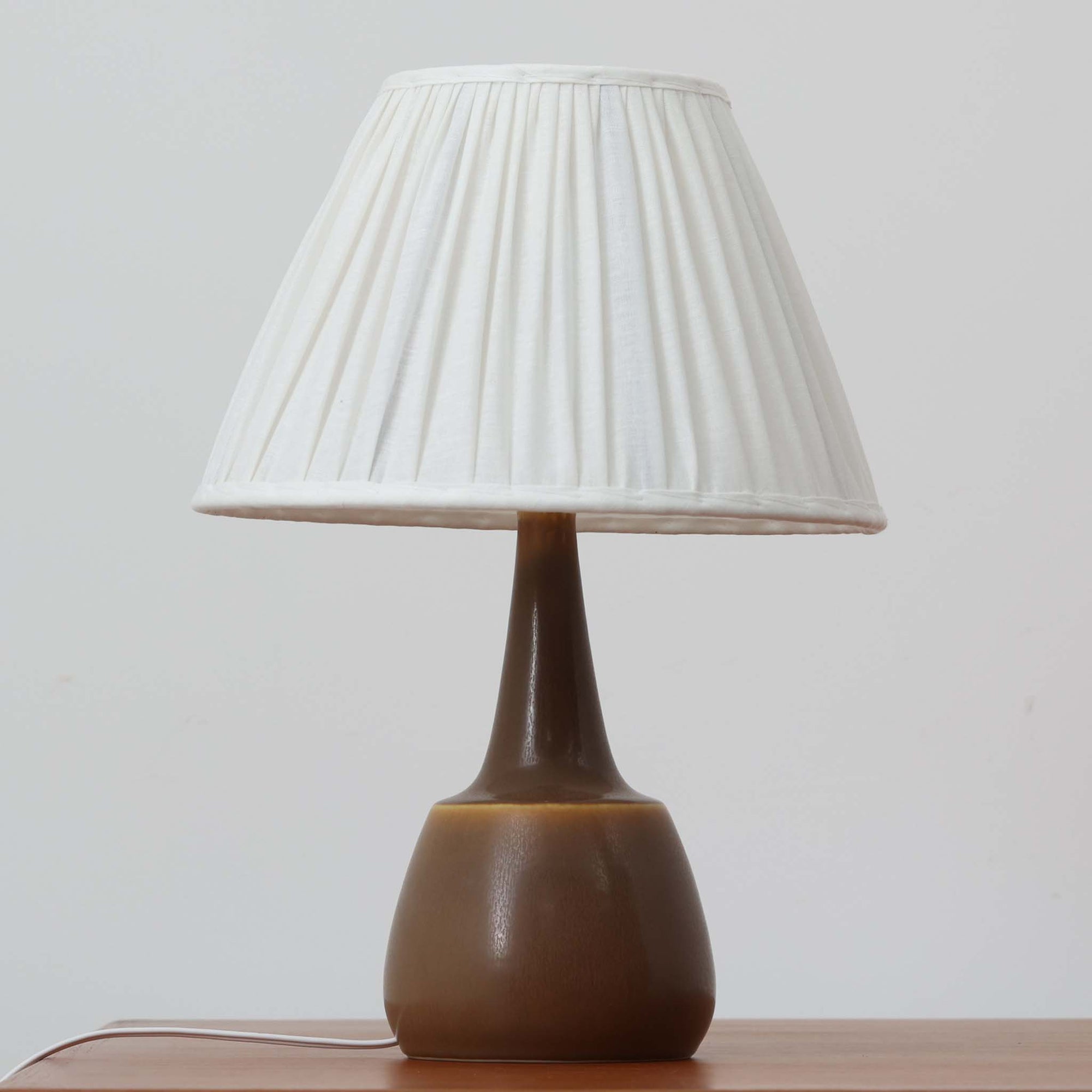 Per Lindemann SchmidtがPalshusのために手掛けたモデル「PL-5 SL 2」は、均整の取れたシルエットと滑らかに焼き締められたマットブラウンの陶肌が静かな存在感を放つテーブルランプで、首元にほんのり入る明るい釉薬のグラデーションが柔らかな表情を添える北欧ビンテージ品です。プリーツシェード越しに広がる落ち着いた灯りはリビングやベッドサイドに心地よい温もりをもたらし、シンプルながら洗練された造形美と経年で深まった風合いを長く楽しめる一台です。