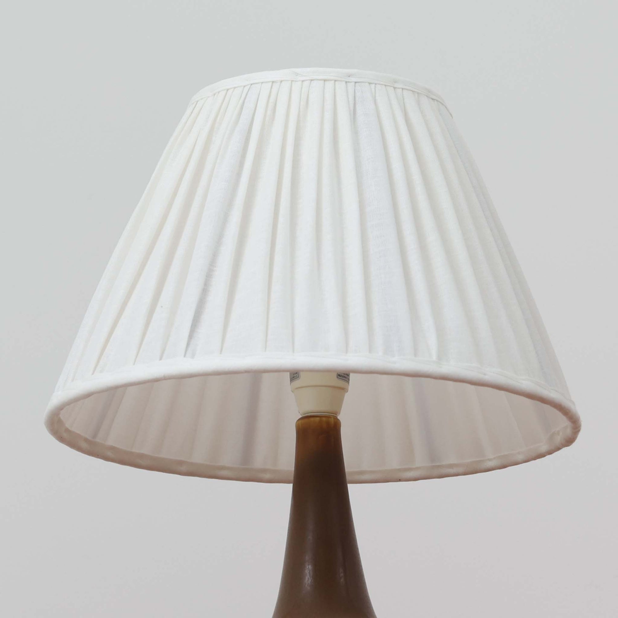 Per Lindemann SchmidtがPalshusのために手掛けたモデル「PL-5 SL 2」は、均整の取れたシルエットと滑らかに焼き締められたマットブラウンの陶肌が静かな存在感を放つテーブルランプで、首元にほんのり入る明るい釉薬のグラデーションが柔らかな表情を添える北欧ビンテージ品です。プリーツシェード越しに広がる落ち着いた灯りはリビングやベッドサイドに心地よい温もりをもたらし、シンプルながら洗練された造形美と経年で深まった風合いを長く楽しめる一台です。