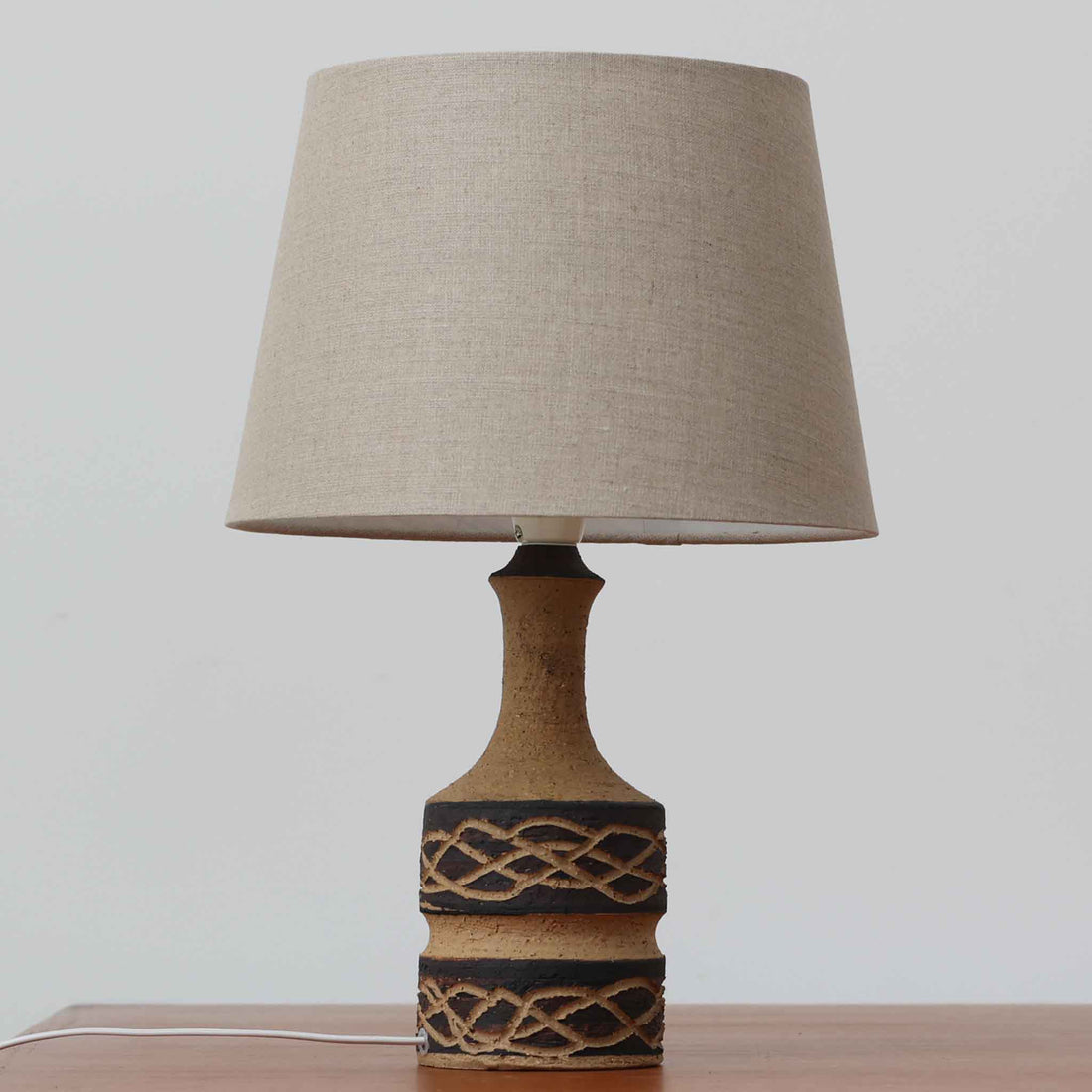 Jette HelleroeがAxellaのために手掛けたこちらのテーブルランプは、砂質の陶肌を生かした素朴で温かみのある質感と、ベース部分にぐるりと施された幾何学的なレリーフ装飾が印象的で、手仕事の風合いが際立つ北欧ビンテージ品です。すらりと立ち上がるシルエットが軽やかなバランスをつくり、リネンシェードを透ける柔らかな灯りがリビングやベッドサイドに落ち着いた雰囲気を添え、経年で深まった土味と装飾の陰影が一点物ならではの魅力を一層引き立てています。
