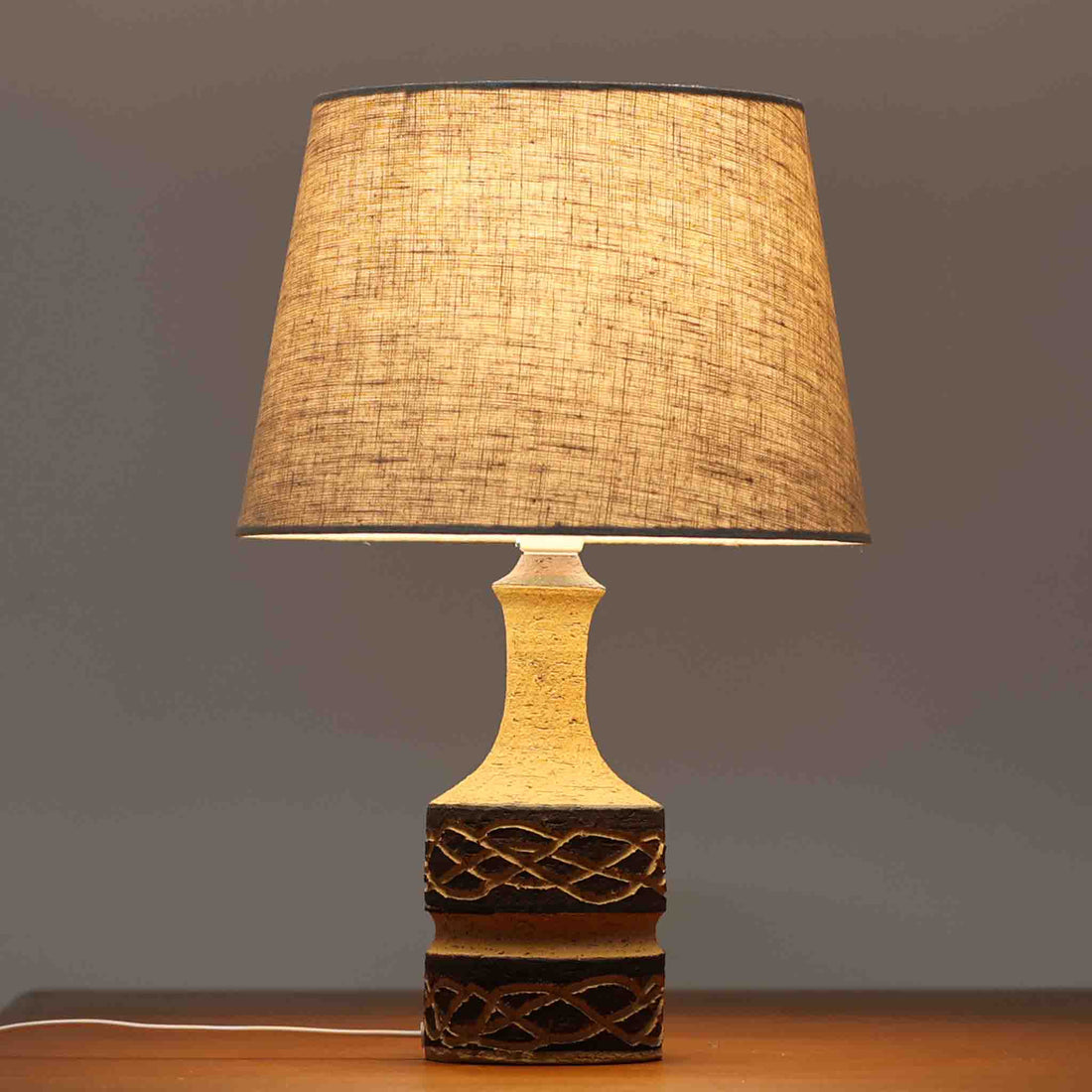 Jette HelleroeがAxellaのために手掛けたこちらのテーブルランプは、砂質の陶肌を生かした素朴で温かみのある質感と、ベース部分にぐるりと施された幾何学的なレリーフ装飾が印象的で、手仕事の風合いが際立つ北欧ビンテージ品です。すらりと立ち上がるシルエットが軽やかなバランスをつくり、リネンシェードを透ける柔らかな灯りがリビングやベッドサイドに落ち着いた雰囲気を添え、経年で深まった土味と装飾の陰影が一点物ならではの魅力を一層引き立てています。