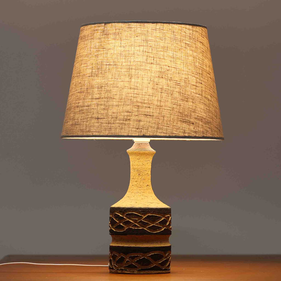 Jette HelleroeがAxellaのために手掛けたこちらのテーブルランプは、砂質の陶肌を生かした素朴で温かみのある質感と、ベース部分にぐるりと施された幾何学的なレリーフ装飾が印象的で、手仕事の風合いが際立つ北欧ビンテージ品です。すらりと立ち上がるシルエットが軽やかなバランスをつくり、リネンシェードを透ける柔らかな灯りがリビングやベッドサイドに落ち着いた雰囲気を添え、経年で深まった土味と装飾の陰影が一点物ならではの魅力を一層引き立てています。