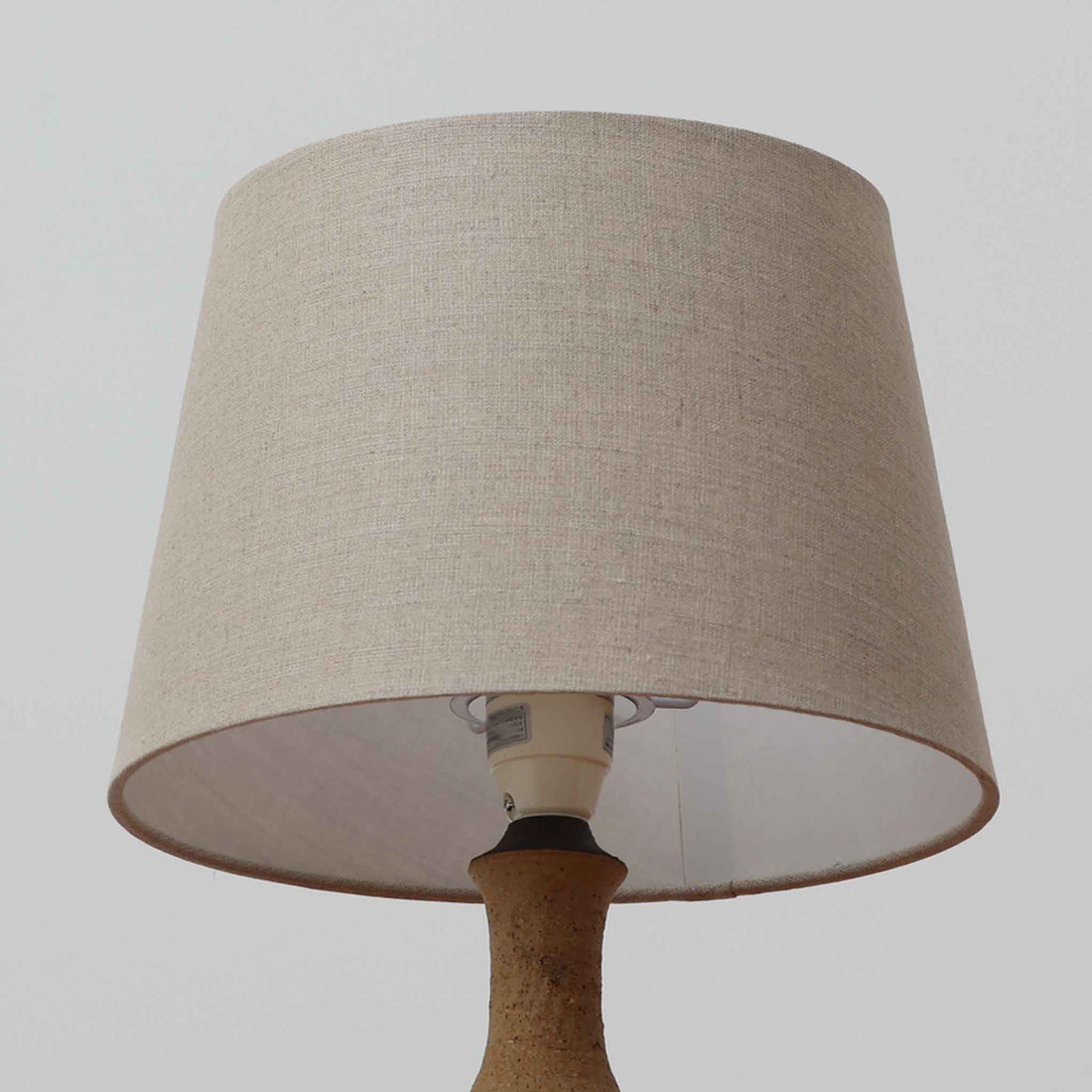 Jette HelleroeがAxellaのために手掛けたこちらのテーブルランプは、砂質の陶肌を生かした素朴で温かみのある質感と、ベース部分にぐるりと施された幾何学的なレリーフ装飾が印象的で、手仕事の風合いが際立つ北欧ビンテージ品です。すらりと立ち上がるシルエットが軽やかなバランスをつくり、リネンシェードを透ける柔らかな灯りがリビングやベッドサイドに落ち着いた雰囲気を添え、経年で深まった土味と装飾の陰影が一点物ならではの魅力を一層引き立てています。