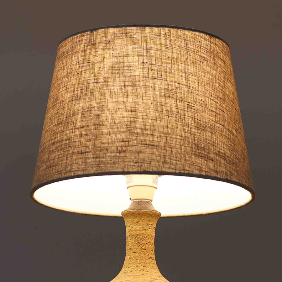 Jette HelleroeがAxellaのために手掛けたこちらのテーブルランプは、砂質の陶肌を生かした素朴で温かみのある質感と、ベース部分にぐるりと施された幾何学的なレリーフ装飾が印象的で、手仕事の風合いが際立つ北欧ビンテージ品です。すらりと立ち上がるシルエットが軽やかなバランスをつくり、リネンシェードを透ける柔らかな灯りがリビングやベッドサイドに落ち着いた雰囲気を添え、経年で深まった土味と装飾の陰影が一点物ならではの魅力を一層引き立てています。