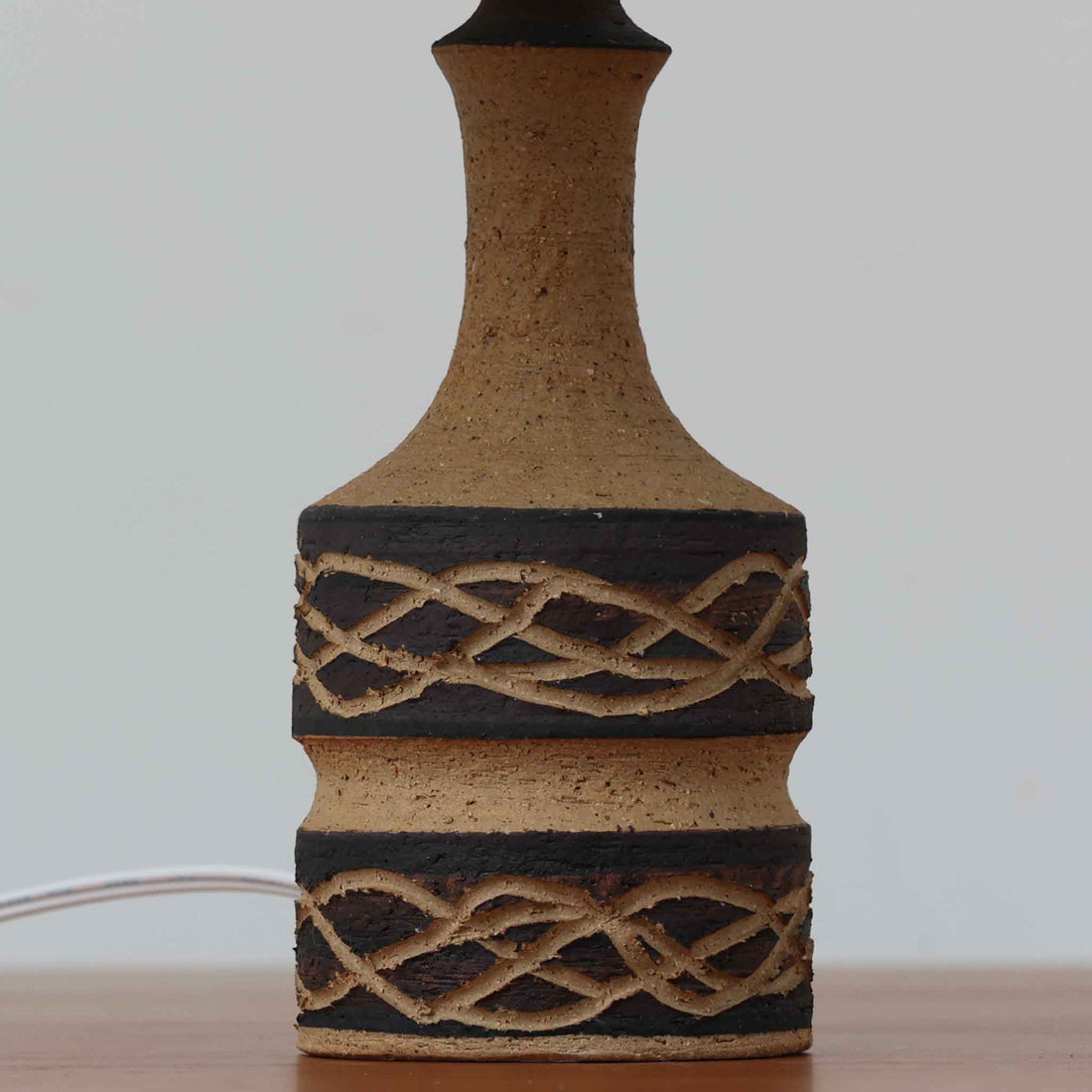 Jette HelleroeがAxellaのために手掛けたこちらのテーブルランプは、砂質の陶肌を生かした素朴で温かみのある質感と、ベース部分にぐるりと施された幾何学的なレリーフ装飾が印象的で、手仕事の風合いが際立つ北欧ビンテージ品です。すらりと立ち上がるシルエットが軽やかなバランスをつくり、リネンシェードを透ける柔らかな灯りがリビングやベッドサイドに落ち着いた雰囲気を添え、経年で深まった土味と装飾の陰影が一点物ならではの魅力を一層引き立てています。