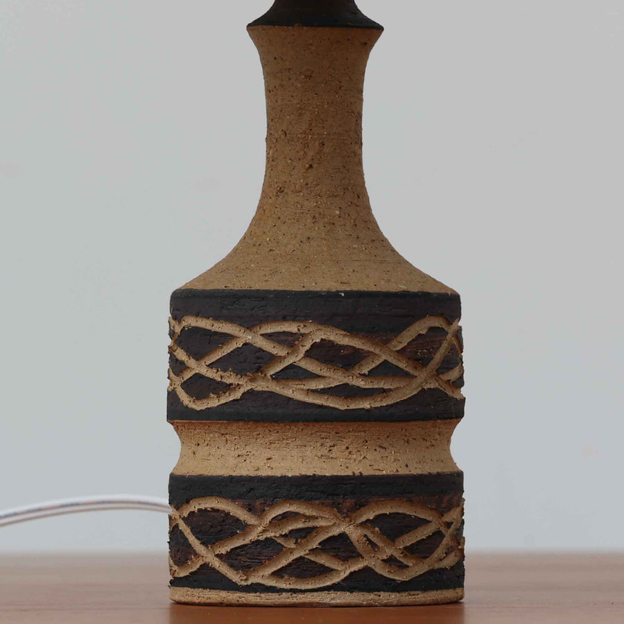 Jette HelleroeがAxellaのために手掛けたこちらのテーブルランプは、砂質の陶肌を生かした素朴で温かみのある質感と、ベース部分にぐるりと施された幾何学的なレリーフ装飾が印象的で、手仕事の風合いが際立つ北欧ビンテージ品です。すらりと立ち上がるシルエットが軽やかなバランスをつくり、リネンシェードを透ける柔らかな灯りがリビングやベッドサイドに落ち着いた雰囲気を添え、経年で深まった土味と装飾の陰影が一点物ならではの魅力を一層引き立てています。
