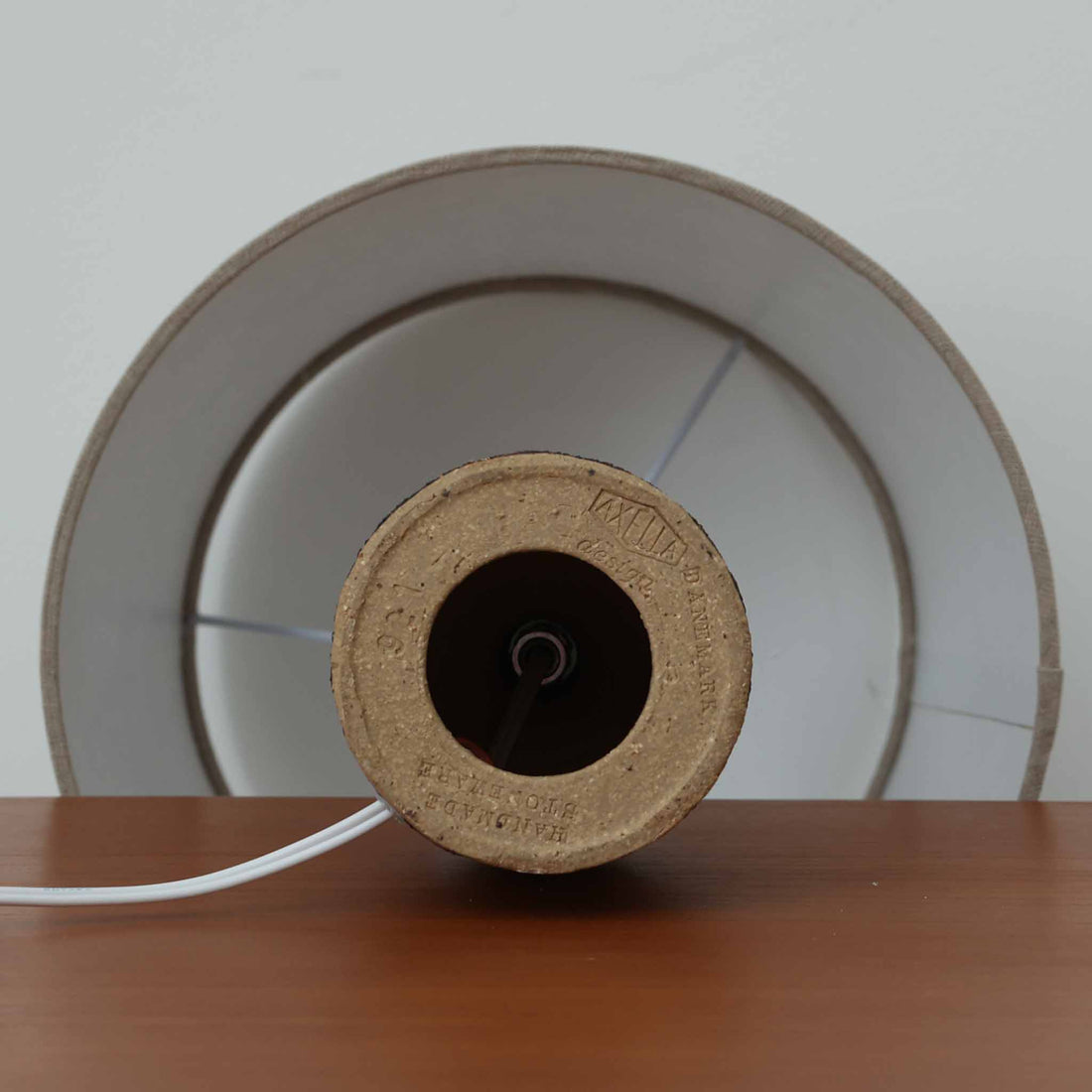 Jette HelleroeがAxellaのために手掛けたこちらのテーブルランプは、砂質の陶肌を生かした素朴で温かみのある質感と、ベース部分にぐるりと施された幾何学的なレリーフ装飾が印象的で、手仕事の風合いが際立つ北欧ビンテージ品です。すらりと立ち上がるシルエットが軽やかなバランスをつくり、リネンシェードを透ける柔らかな灯りがリビングやベッドサイドに落ち着いた雰囲気を添え、経年で深まった土味と装飾の陰影が一点物ならではの魅力を一層引き立てています。