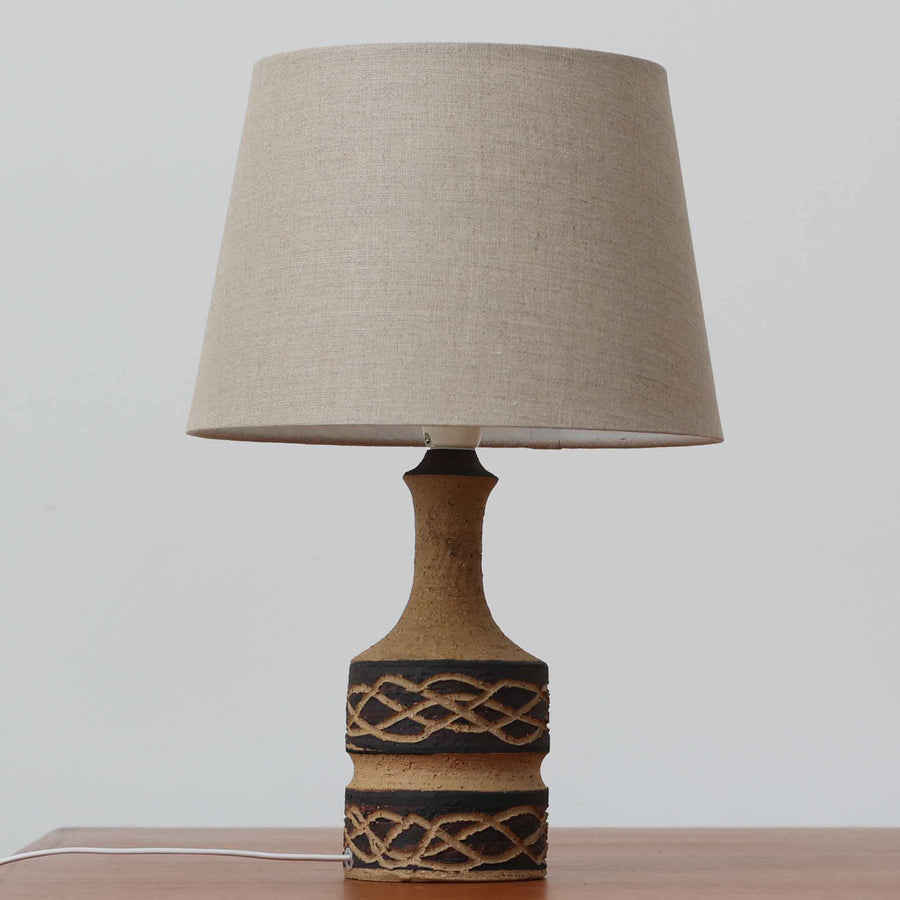 Jette HelleroeがAxellaのために手掛けたこちらのテーブルランプは、砂質の陶肌を生かした素朴で温かみのある質感と、ベース部分にぐるりと施された幾何学的なレリーフ装飾が印象的で、手仕事の風合いが際立つ北欧ビンテージ品です。すらりと立ち上がるシルエットが軽やかなバランスをつくり、リネンシェードを透ける柔らかな灯りがリビングやベッドサイドに落ち着いた雰囲気を添え、経年で深まった土味と装飾の陰影が一点物ならではの魅力を一層引き立てています。