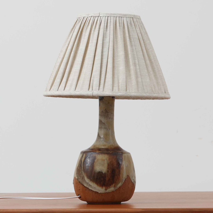 Axella製のこちらのテーブルランプは、素地を生かした陶土の質感に流れるような釉薬のグラデーションが重なり合う有機的な表情が魅力で、ぽってりとした丸みのあるフォルムと細く立ち上がる首部の対比が北欧らしい温かみのある佇まいを生み出すビンテージ品です。手仕事ならではの不均一な釉薬の景色が一点物の存在感を与え、リネンシェードからこぼれる柔らかな灯りがリビングや寝室に落ち着いた雰囲気を添え、経年で深まった陶肌の風合いが空間に自然なアクセントを加えてくれます。