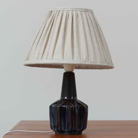 Einar JohansenがSoholm Keramikのために手掛けたモデル「1052」は、濃淡のある深いブルーグラデーションと面取りを施した立体的なフォルムが美しく、上部のマットな質感とのコントラストが上品な存在感を生む北欧ビンテージのテーブルランプです。柔らかく広がるリネンシェードの灯りが陰影豊かな陶肌をやさしく照らし、リビングや寝室のアクセントとして取り入れやすく、経年で深まった釉薬の表情が一点物ならではの味わいを引き立てています。