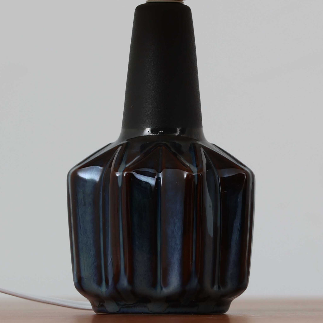 Einar JohansenがSoholm Keramikのために手掛けたモデル「1052」は、濃淡のある深いブルーグラデーションと面取りを施した立体的なフォルムが美しく、上部のマットな質感とのコントラストが上品な存在感を生む北欧ビンテージのテーブルランプです。柔らかく広がるリネンシェードの灯りが陰影豊かな陶肌をやさしく照らし、リビングや寝室のアクセントとして取り入れやすく、経年で深まった釉薬の表情が一点物ならではの味わいを引き立てています。