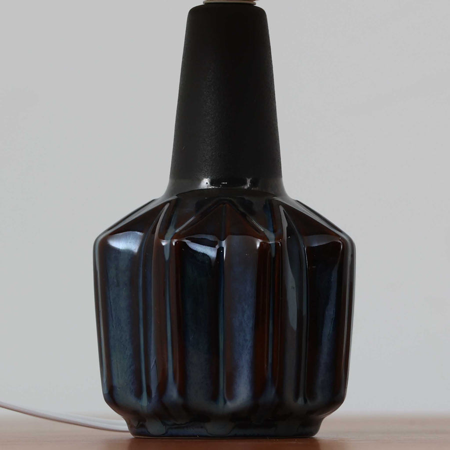 Einar JohansenがSoholm Keramikのために手掛けたモデル「1052」は、濃淡のある深いブルーグラデーションと面取りを施した立体的なフォルムが美しく、上部のマットな質感とのコントラストが上品な存在感を生む北欧ビンテージのテーブルランプです。柔らかく広がるリネンシェードの灯りが陰影豊かな陶肌をやさしく照らし、リビングや寝室のアクセントとして取り入れやすく、経年で深まった釉薬の表情が一点物ならではの味わいを引き立てています。