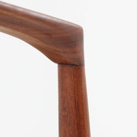 Kai Kristiansenがデザインし、Magnus Olesenが製作したTrojaアームチェアです。アフロモシア材の滑らかな木肌と深みのある色合いが美しく、背からアームへと一体で削り出されたフレームが身体をやさしく包み込みます。わずかに湾曲した笠木や細身の脚部が軽快な印象を与え、立ち座りの動作も自然。張り替え済みの座面は快適な掛け心地を備え、ダイニングや書斎空間に北欧ビンテージならではの気品と造形美をもたらす一脚です。ロゴは、デンマークの家具販売店（小売店）である Brdr. Ellehuus（エルフース兄弟店） のものです。