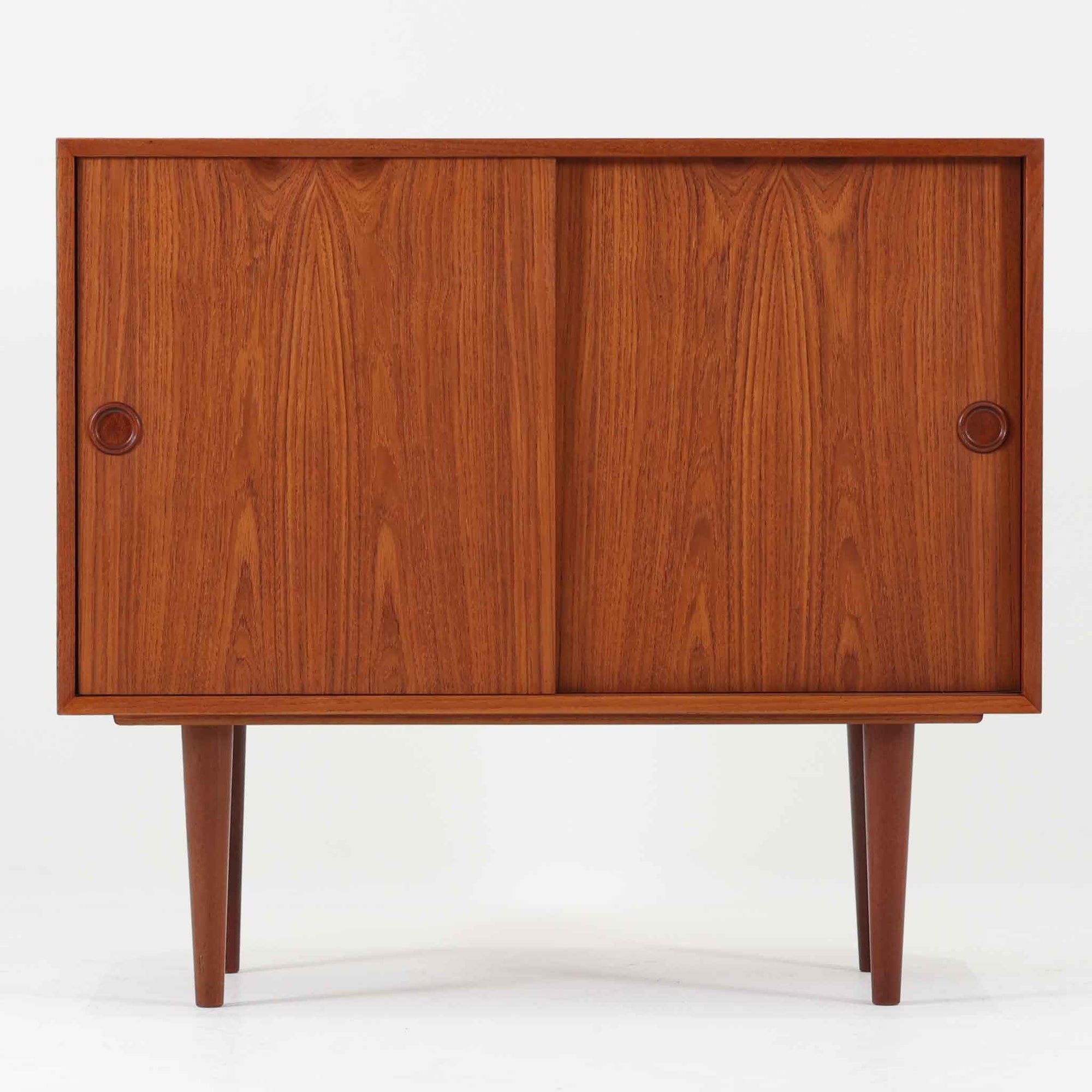 Kai Kristiansenがデザインし、Feldballes Mobelfabrikが製作した幅86cmの小ぶりなボードです。チーク材の美しい木目を活かした端正な箱型フォルムに、丸い引き手付きの引き戸がアクセントを添えます。内部には棚とトレー収納を備え、書類や小物を整理しやすい実用的な構造。すらりと伸びる脚部が軽やかさを演出し、限られた空間にも取り入れやすいサイズ感です。経年により深みを増した木肌が、北欧ビンテージならではの温もりと存在感を感じさせます。