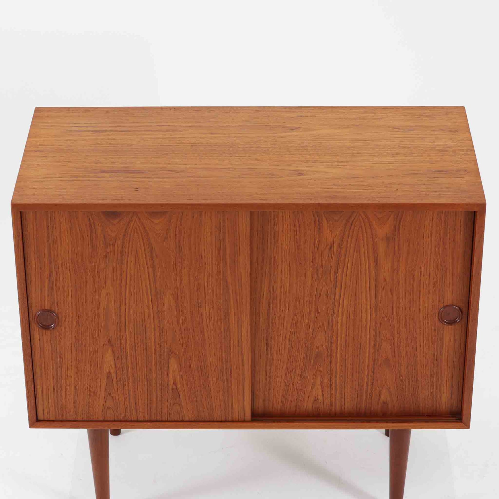 Kai Kristiansenがデザインし、Feldballes Mobelfabrikが製作した幅86cmの小ぶりなボードです。チーク材の美しい木目を活かした端正な箱型フォルムに、丸い引き手付きの引き戸がアクセントを添えます。内部には棚とトレー収納を備え、書類や小物を整理しやすい実用的な構造。すらりと伸びる脚部が軽やかさを演出し、限られた空間にも取り入れやすいサイズ感です。経年により深みを増した木肌が、北欧ビンテージならではの温もりと存在感を感じさせます。