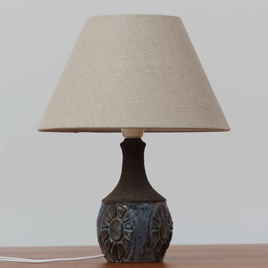 Chris HaslevがJetiのために手掛けたこちらのテーブルランプは、太陽を思わせるレリーフ模様をまとった陶器ベースと、素朴な土肌と釉薬の濃淡が織りなす豊かな表情が魅力で、コンパクトながら存在感を放つ北欧ビンテージ品です。上部のマットな質感が造形を引き締め、リネンシェード越しに広がるあたたかな灯りが空間をやさしく包み込み、リビングやベッドサイドのアクセントとして取り入れやすい一台に仕上がっています。