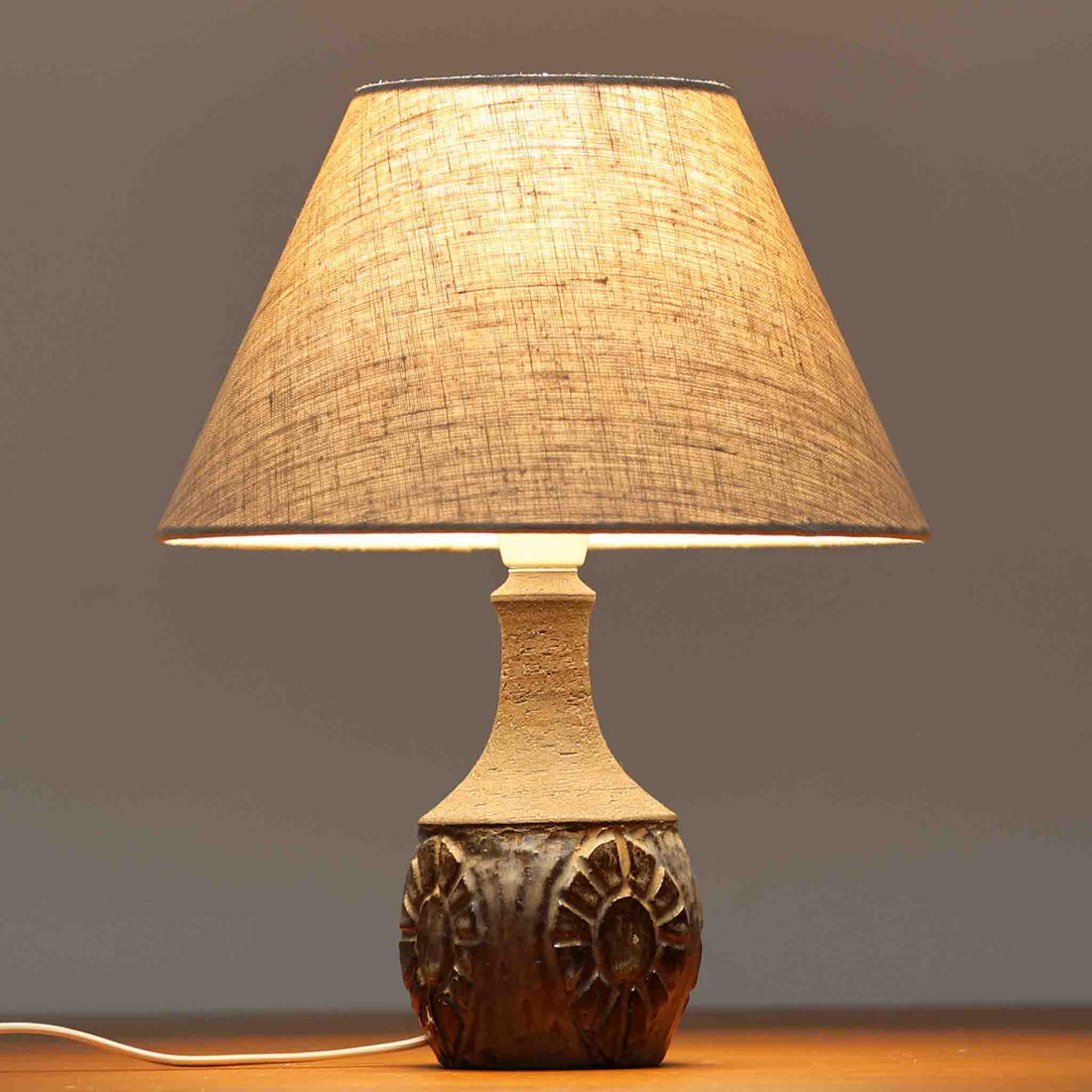 Chris HaslevがJetiのために手掛けたこちらのテーブルランプは、太陽を思わせるレリーフ模様をまとった陶器ベースと、素朴な土肌と釉薬の濃淡が織りなす豊かな表情が魅力で、コンパクトながら存在感を放つ北欧ビンテージ品です。上部のマットな質感が造形を引き締め、リネンシェード越しに広がるあたたかな灯りが空間をやさしく包み込み、リビングやベッドサイドのアクセントとして取り入れやすい一台に仕上がっています。