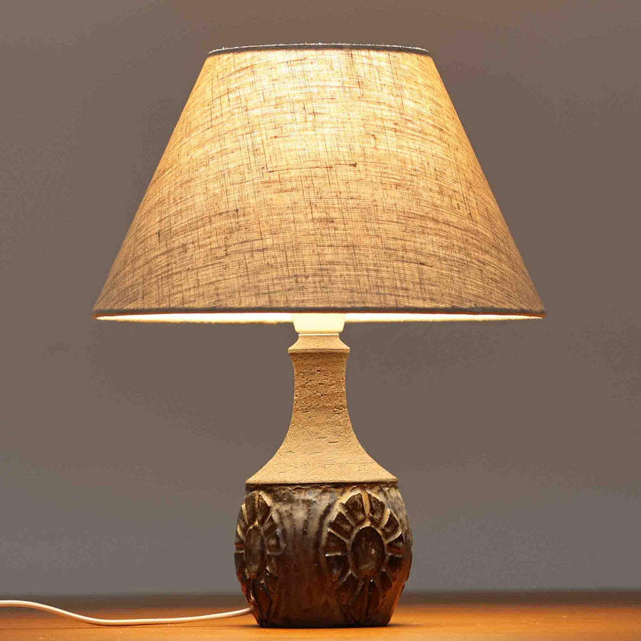 Chris HaslevがJetiのために手掛けたこちらのテーブルランプは、太陽を思わせるレリーフ模様をまとった陶器ベースと、素朴な土肌と釉薬の濃淡が織りなす豊かな表情が魅力で、コンパクトながら存在感を放つ北欧ビンテージ品です。上部のマットな質感が造形を引き締め、リネンシェード越しに広がるあたたかな灯りが空間をやさしく包み込み、リビングやベッドサイドのアクセントとして取り入れやすい一台に仕上がっています。