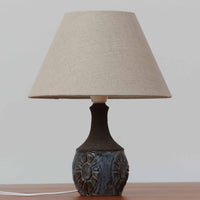 Chris HaslevがJetiのために手掛けたこちらのテーブルランプは、太陽を思わせるレリーフ模様をまとった陶器ベースと、素朴な土肌と釉薬の濃淡が織りなす豊かな表情が魅力で、コンパクトながら存在感を放つ北欧ビンテージ品です。上部のマットな質感が造形を引き締め、リネンシェード越しに広がるあたたかな灯りが空間をやさしく包み込み、リビングやベッドサイドのアクセントとして取り入れやすい一台に仕上がっています。