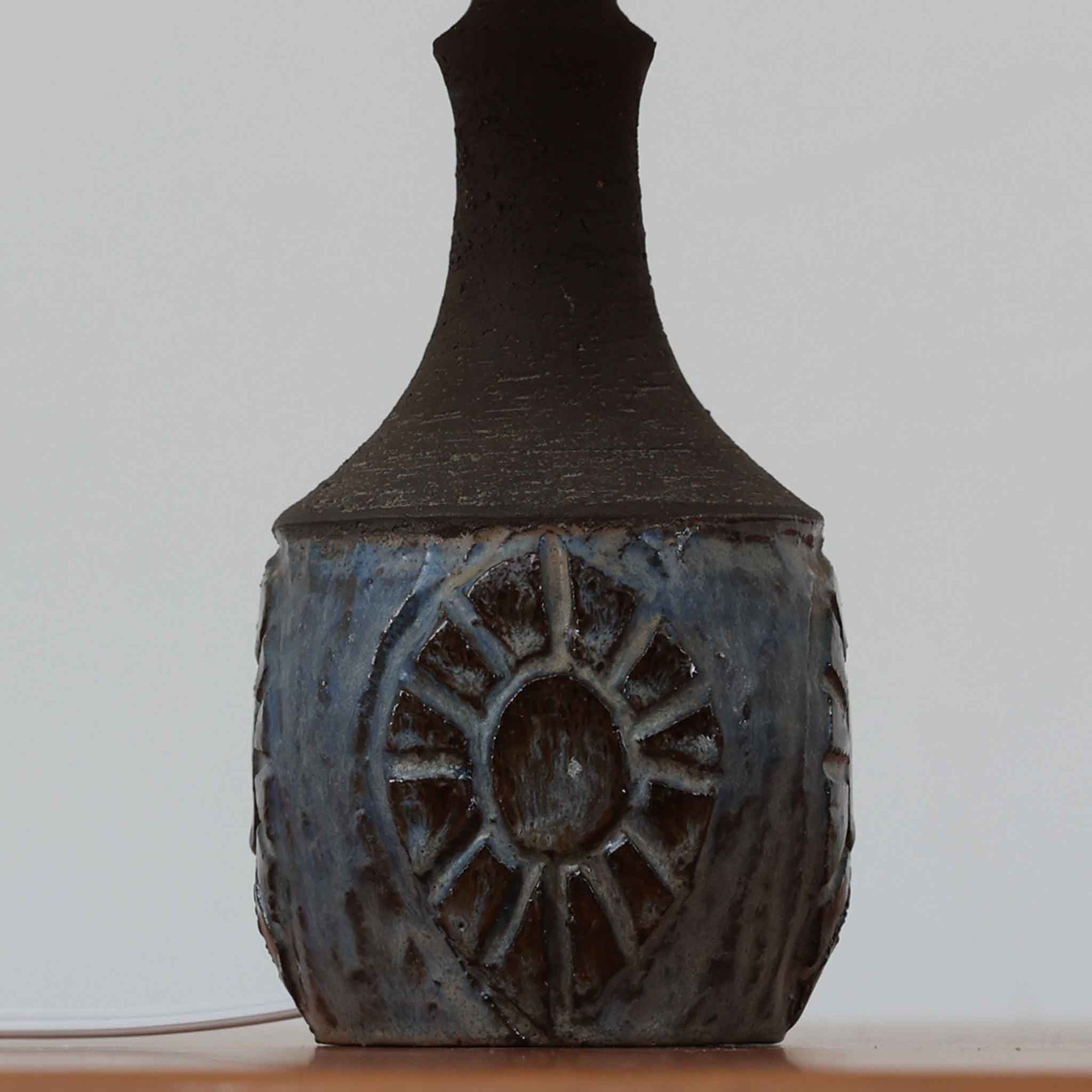 Chris HaslevがJetiのために手掛けたこちらのテーブルランプは、太陽を思わせるレリーフ模様をまとった陶器ベースと、素朴な土肌と釉薬の濃淡が織りなす豊かな表情が魅力で、コンパクトながら存在感を放つ北欧ビンテージ品です。上部のマットな質感が造形を引き締め、リネンシェード越しに広がるあたたかな灯りが空間をやさしく包み込み、リビングやベッドサイドのアクセントとして取り入れやすい一台に仕上がっています。