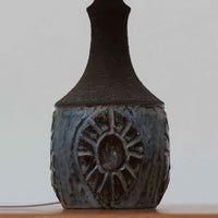 Chris HaslevがJetiのために手掛けたこちらのテーブルランプは、太陽を思わせるレリーフ模様をまとった陶器ベースと、素朴な土肌と釉薬の濃淡が織りなす豊かな表情が魅力で、コンパクトながら存在感を放つ北欧ビンテージ品です。上部のマットな質感が造形を引き締め、リネンシェード越しに広がるあたたかな灯りが空間をやさしく包み込み、リビングやベッドサイドのアクセントとして取り入れやすい一台に仕上がっています。