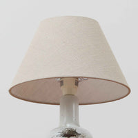 Nils Thorsson（ニルス・トールソン）によるRoyal Copenhagen（ロイヤルコペンハーゲン）の「DIANA」シリーズのテーブルランプは、白磁に繊細な草花や鳥の情景が描かれた静謐な佇まいが魅力で、淡い釉調が経年によりさらに深みを増したビンテージならではの味わいを感じさせます。柔らかく丸みを帯びたベースから伸びるすっきりとした首部のプロポーションが美しく、リネンシェードを通した穏やかな灯りが空間に上質な落ち着きを添えます。高さ32cmとコンパクトで取り入れやすく、ベッドサイドや読書灯としても活躍する、北欧陶芸の上品さが詰まった一台です。