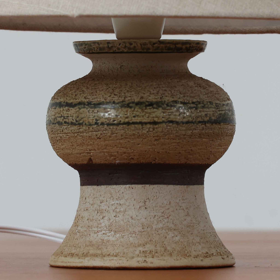 Axella製のこちらのテーブルランプは、素朴な陶土の質感と水平に重ねられた釉薬のグラデーションが生む落ち着いた佇まいが魅力で、ふくらみのある曲線的なベースと小ぶりなサイズ感が空間にさりげない温かみを添える北欧ビンテージ品です。リネンシェード越しに漏れる柔らかな灯りが陶肌の陰影をやさしく引き立て、デスクやベッドサイドなどにも取り入れやすく、経年で深まった素材の風合いが一点物ならではの味わいを感じさせてくれます。