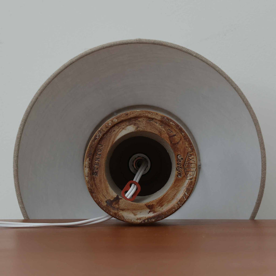 Axella製のこちらのテーブルランプは、素朴な陶土の質感と水平に重ねられた釉薬のグラデーションが生む落ち着いた佇まいが魅力で、ふくらみのある曲線的なベースと小ぶりなサイズ感が空間にさりげない温かみを添える北欧ビンテージ品です。リネンシェード越しに漏れる柔らかな灯りが陶肌の陰影をやさしく引き立て、デスクやベッドサイドなどにも取り入れやすく、経年で深まった素材の風合いが一点物ならではの味わいを感じさせてくれます。