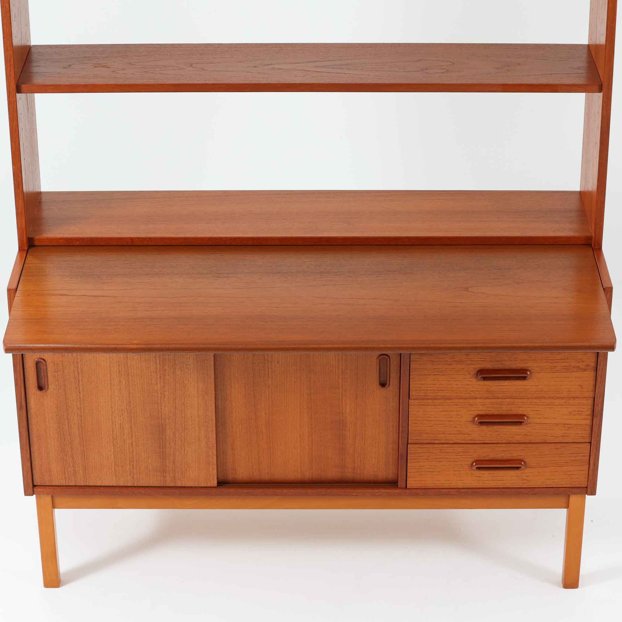 チーク材とビーチ材を組み合わせた北欧ビンテージらしい佇まいが魅力のディスプレイシェルフで、直線的で端正なフレーム構成と、木目の表情を活かした温かみのある質感が空間に落ち着きをもたらします。上部のオープン棚は器や書籍、オブジェの展示に適しており、下部には引き戸収納と引き出しを備えることで、見せる収納と隠す収納をバランス良く両立しています。脚付き構造により圧迫感が少なく、リビングやダイニング、書斎など幅広いシーンで活躍し、経年変化による色味の深まりも楽しめる一台です。