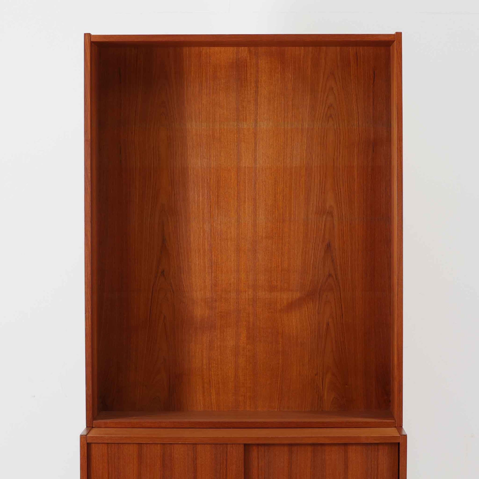 Asljunga Mobelfabrikによるディスプレイシェルフは、チーク材とビーチ材を組み合わせた端正な佇まいが魅力の北欧ビンテージ家具です。上段はオープンシェルフ構造で、棚板を活かした見せる収納が楽しめ、下段には引き戸式の収納部を備えることで実用性も確保されています。直線的で無駄のないフォルムに、掘り込み式の取っ手や脚部の軽やかなバランスが北欧らしい品の良さを添え、幅79cmの程よいサイズ感はリビングやダイニング、書斎など幅広い空間に取り入れやすく、経年変化によって深みを増した木肌と丁寧な造りが静かな存在感を放つ一台です。