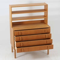 B. FridhagenがBodaforsのために手掛けたディスプレイシェルフは、端正なプロポーションと実用性を兼ね備えたスウェーデン製ビンテージ家具です。上部は抜け感のあるオープン棚構造で、器や書籍、オブジェなどを軽やかに飾ることができ、下部には4段の引き出しを備え、日常使いの収納にも対応します。オーク材ならではの明るく上品な木肌と、直線を基調とした構成が北欧らしい落ち着いた印象を生み、空間をすっきりとまとめてくれます。幅90cm、高さ122cmの小ぶりなサイズ感で、ダイニングやリビング、書斎など幅広いシーンに取り入れやすく、経年変化による木の表情も楽しめる一台です。