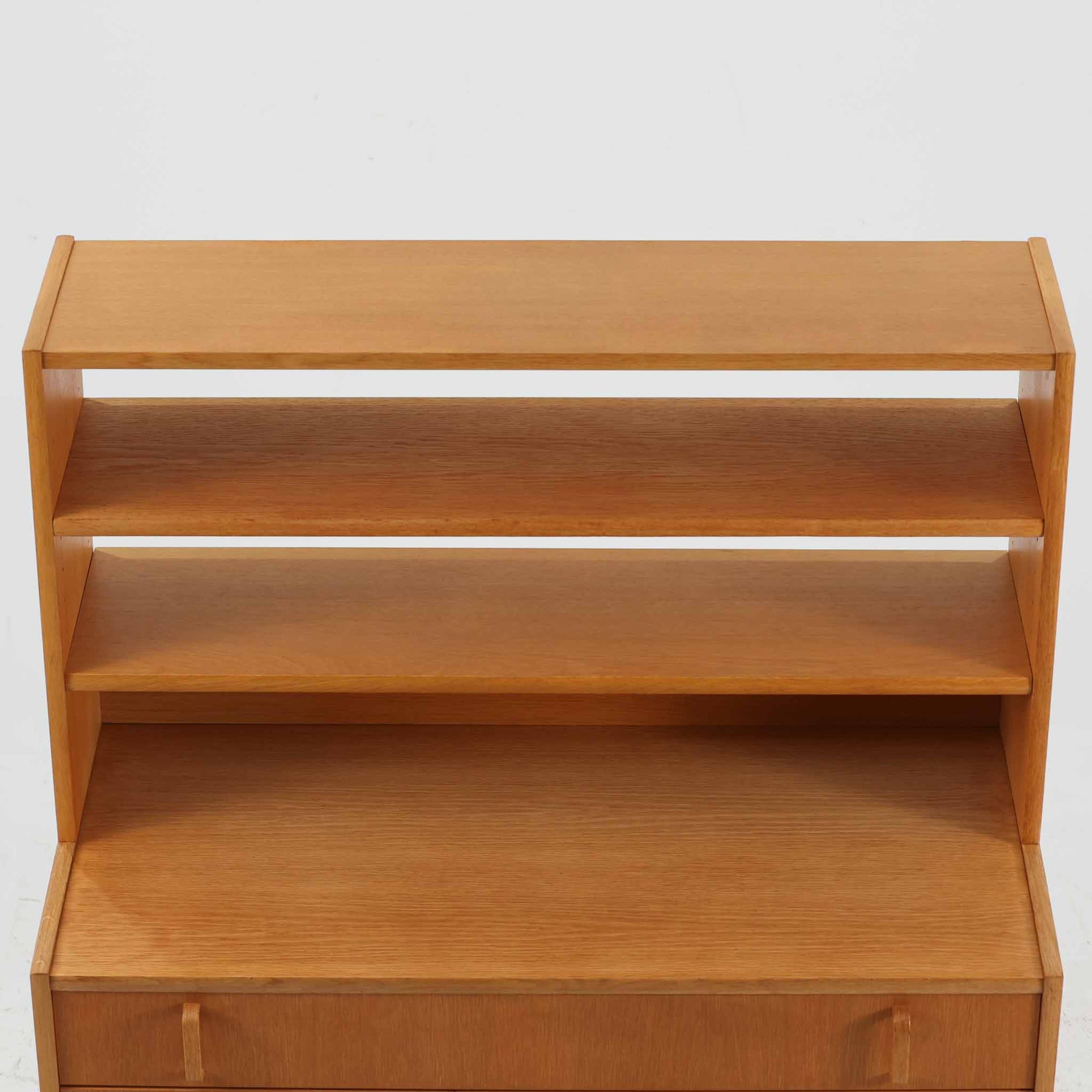 B. FridhagenがBodaforsのために手掛けたディスプレイシェルフは、端正なプロポーションと実用性を兼ね備えたスウェーデン製ビンテージ家具です。上部は抜け感のあるオープン棚構造で、器や書籍、オブジェなどを軽やかに飾ることができ、下部には4段の引き出しを備え、日常使いの収納にも対応します。オーク材ならではの明るく上品な木肌と、直線を基調とした構成が北欧らしい落ち着いた印象を生み、空間をすっきりとまとめてくれます。幅90cm、高さ122cmの小ぶりなサイズ感で、ダイニングやリビング、書斎など幅広いシーンに取り入れやすく、経年変化による木の表情も楽しめる一台です。