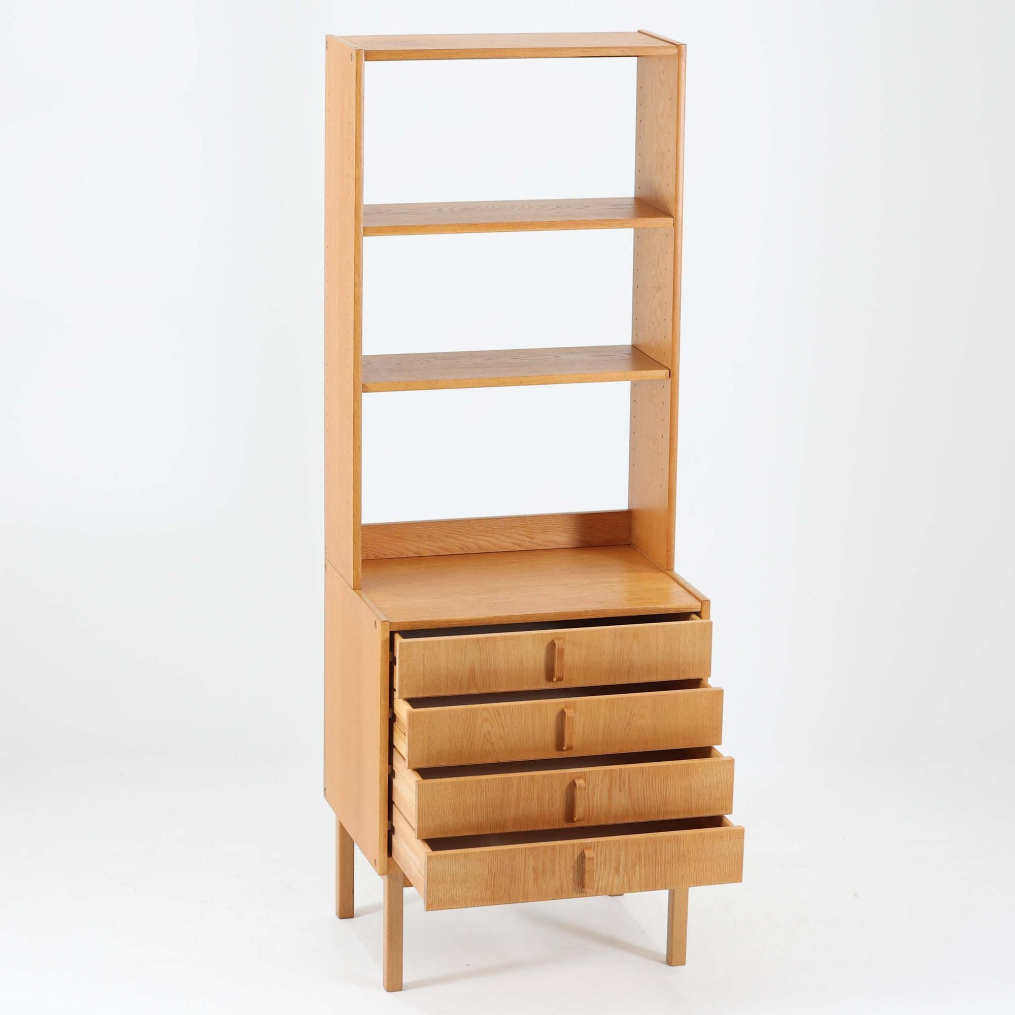B. Fridhagenがデザインし、Bodafors社により1963年に製作されたスリムシェルフです。オーク材のやわらかな木目と明るい色合いが空間に温もりをもたらし、上部のオープン棚と下部の4段引き出しを備えた実用的な構成が魅力。幅63cmのすっきりとしたサイズで、書斎やリビングの一角にも取り入れやすい一台です。直線的で端正なフォルムと丁寧なつくりから、北欧ビンテージならではの機能美と経年の味わいを感じられる逸品です。