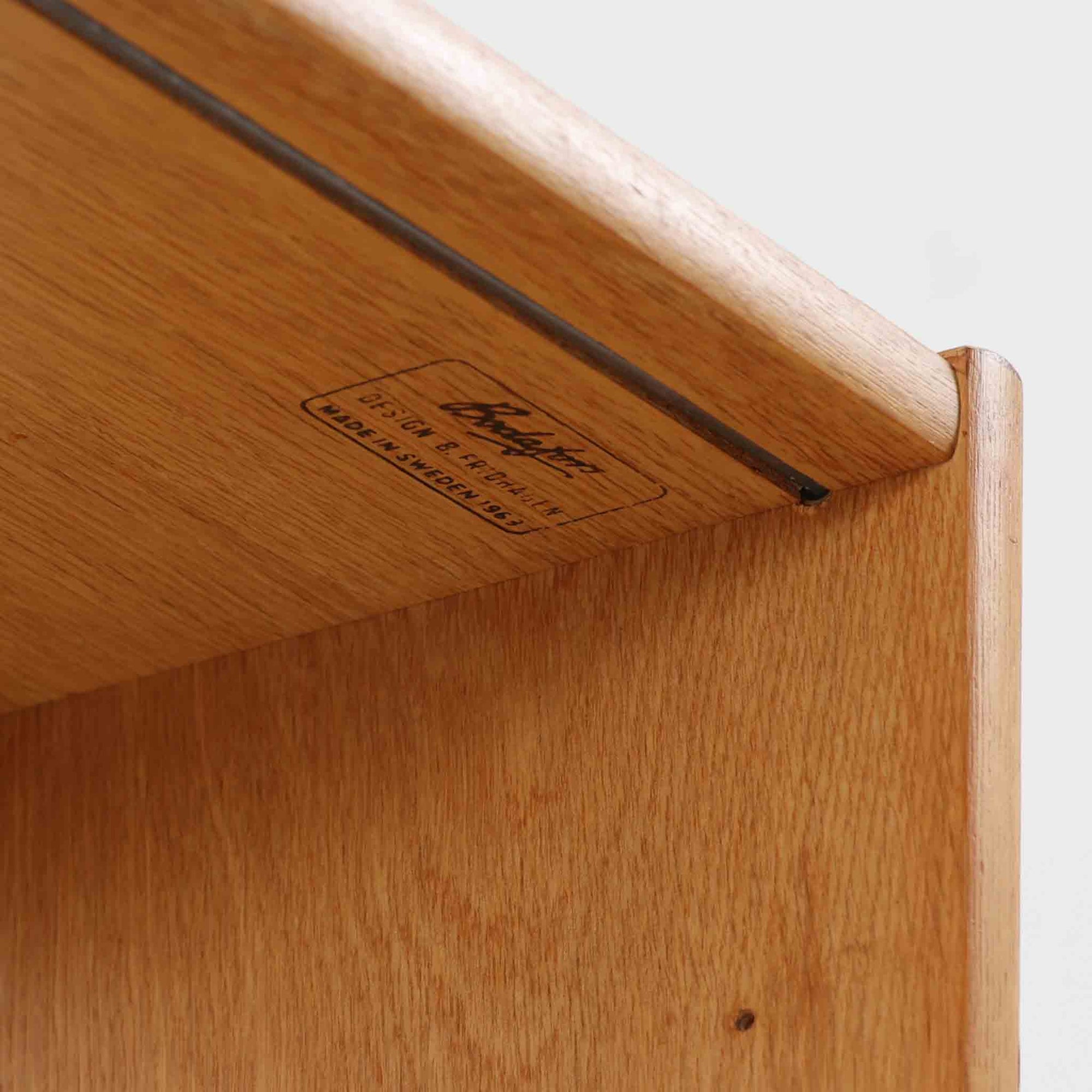 B. Fridhagenがデザインし、Bodafors社により1963年に製作されたスリムシェルフです。オーク材のやわらかな木目と明るい色合いが空間に温もりをもたらし、上部のオープン棚と下部の4段引き出しを備えた実用的な構成が魅力。幅63cmのすっきりとしたサイズで、書斎やリビングの一角にも取り入れやすい一台です。直線的で端正なフォルムと丁寧なつくりから、北欧ビンテージならではの機能美と経年の味わいを感じられる逸品です。