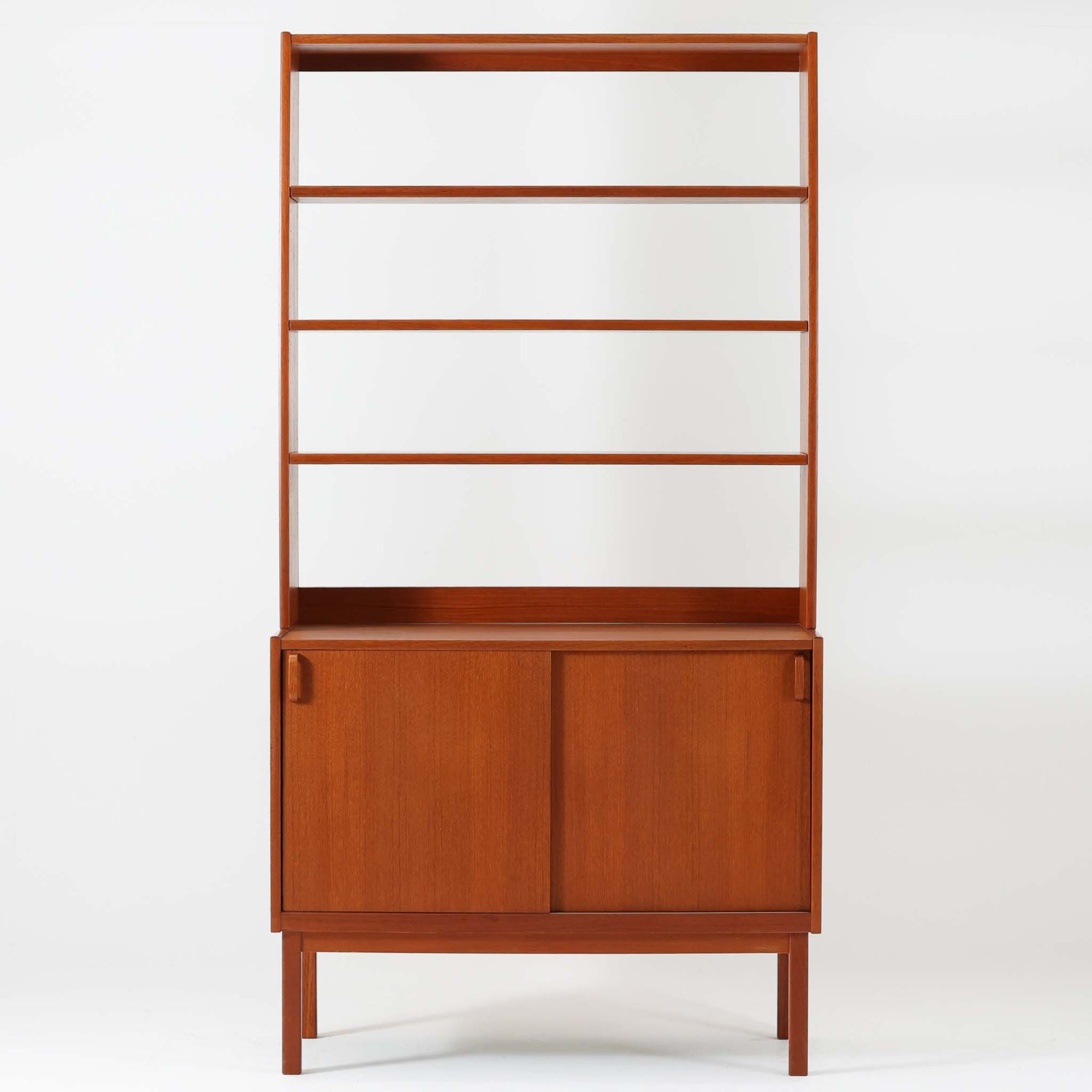 B.フリードホーゲンがデザインし、Bodafors社により製作された北欧ビンテージのディスプレイシェルフです。チーク材の美しい木目を活かした縦に伸びる端正なフレーム構成が印象的で、オープン棚と下部収納を組み合わせた実用性の高いデザインとなっています。上段は抜け感のある棚板構造で、書籍や器、オブジェを軽やかに飾ることができ、下段の引き戸収納は生活感の出やすい物をすっきりと収められます。脚付きによる軽快な佇まいと、経年変化によって深みを増した素材感が、空間に北欧ビンテージならではの落ち着いた存在感を与えます。