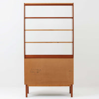 B.フリードホーゲンがデザインし、Bodafors社により製作された北欧ビンテージのディスプレイシェルフです。チーク材の美しい木目を活かした縦に伸びる端正なフレーム構成が印象的で、オープン棚と下部収納を組み合わせた実用性の高いデザインとなっています。上段は抜け感のある棚板構造で、書籍や器、オブジェを軽やかに飾ることができ、下段の引き戸収納は生活感の出やすい物をすっきりと収められます。脚付きによる軽快な佇まいと、経年変化によって深みを増した素材感が、空間に北欧ビンテージならではの落ち着いた存在感を与えます。