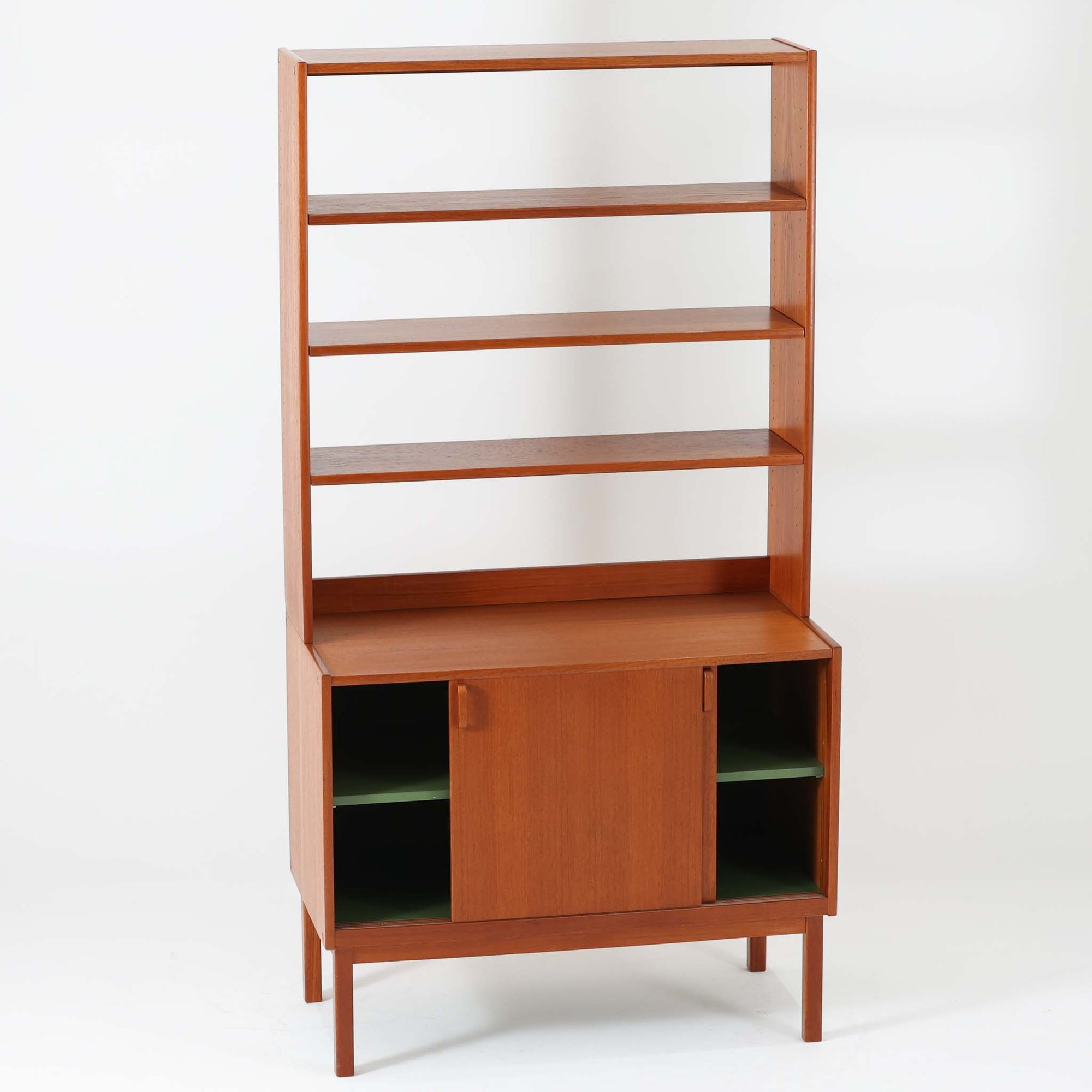B.フリードホーゲンがデザインし、Bodafors社により製作された北欧ビンテージのディスプレイシェルフです。チーク材の美しい木目を活かした縦に伸びる端正なフレーム構成が印象的で、オープン棚と下部収納を組み合わせた実用性の高いデザインとなっています。上段は抜け感のある棚板構造で、書籍や器、オブジェを軽やかに飾ることができ、下段の引き戸収納は生活感の出やすい物をすっきりと収められます。脚付きによる軽快な佇まいと、経年変化によって深みを増した素材感が、空間に北欧ビンテージならではの落ち着いた存在感を与えます。