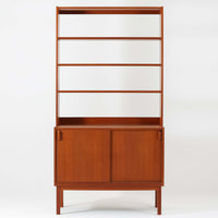 B.フリードホーゲンがデザインし、Bodafors社により製作された北欧ビンテージのディスプレイシェルフです。チーク材の美しい木目を活かした縦に伸びる端正なフレーム構成が印象的で、オープン棚と下部収納を組み合わせた実用性の高いデザインとなっています。上段は抜け感のある棚板構造で、書籍や器、オブジェを軽やかに飾ることができ、下段の引き戸収納は生活感の出やすい物をすっきりと収められます。脚付きによる軽快な佇まいと、経年変化によって深みを増した素材感が、空間に北欧ビンテージならではの落ち着いた存在感を与えます。