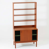 B.フリードホーゲンがデザインし、Bodafors社により製作された北欧ビンテージのディスプレイシェルフです。チーク材の美しい木目を活かした縦に伸びる端正なフレーム構成が印象的で、オープン棚と下部収納を組み合わせた実用性の高いデザインとなっています。上段は抜け感のある棚板構造で、書籍や器、オブジェを軽やかに飾ることができ、下段の引き戸収納は生活感の出やすい物をすっきりと収められます。脚付きによる軽快な佇まいと、経年変化によって深みを増した素材感が、空間に北欧ビンテージならではの落ち着いた存在感を与えます。